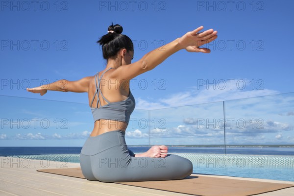 A woman practices yoga by a pool with a stunning ocean view. She sits in lotus pose on a mat, extending her arms with focus and tranquility under a clear blue sky