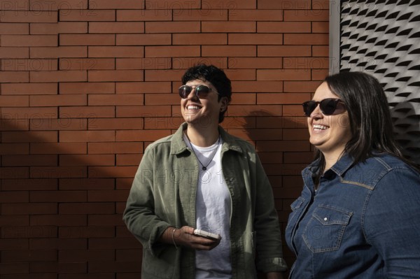 A happy curvy lesbian couple enjoying a sunny day outdoors. They are smiling, wearing casual jackets and sunglasses, embracing love and togetherness against a brick wall backdrop
