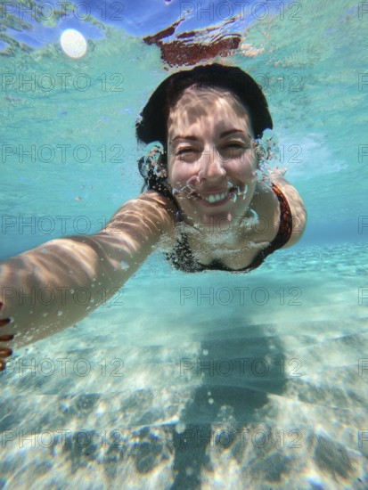 A woman swims underwater in Greece, capturing a selfie. The clear blue water reflects sunlight, creating a playful and joyful atmosphere in the sea