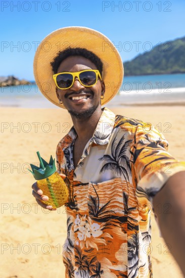 Young man taking a selfie on a tropical beach, wearing sunglasses and a straw hat, holding a pineapple shaped cocktail and smiling