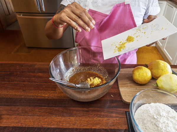 A woman wearing a pink apron prepares lemon cookies in the kitchen. She adds lemon zest to a brown mixture in a glass bowl, with the ingredients arranged on a wooden countertop