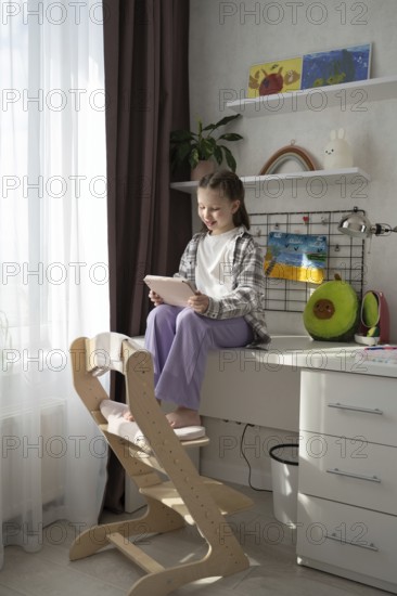 A child sits on a desk, engaged with a tablet, surrounded by vibrant art, plush toys, and houseplants in a warm, well-lit room. The scene evokes creativity and tranquility