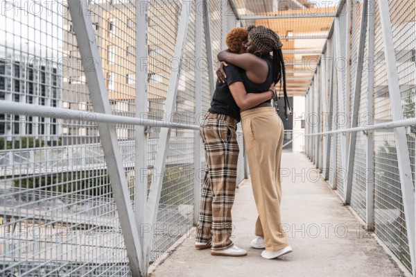 A loving LGBT multiethnic couple embraces on a metal bridge in an urban setting. Their warm hug showcases affection and inclusivity, set against a backdrop of city architecture