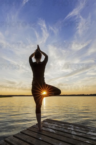 Woman practicing standing posture Vrikshasana, Tree Pose, one legged balancing asana of the medieval hatha yoga on jetty at lake at sunset