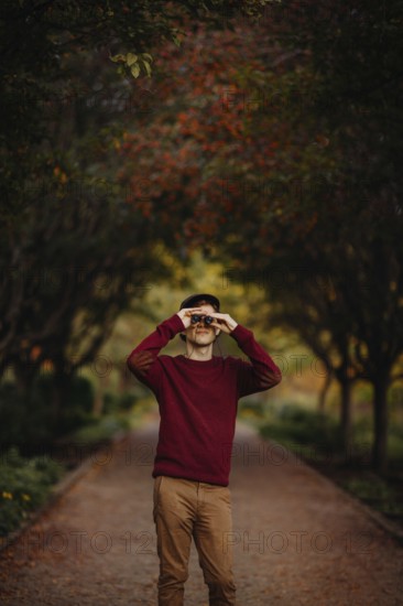 A man in a red sweater looks through binoculars amidst vibrant autumn foliage in a scenic park pathway in Quebec, Canada, capturing the essence of fall's beauty