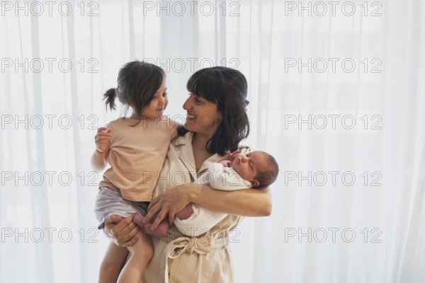 A joyful young family scene with a mother holding her newborn baby and big sister. Both children are in her arms, expressing love and happiness in a bright setting