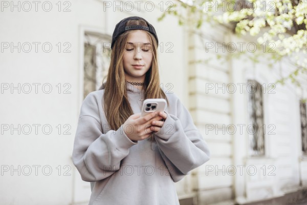 A teenage girl stands outdoors, focused on her smartphone, wearing a casual gray hoodie and a black cap. She is absorbed in her screen, engaging with technology on a sunny day