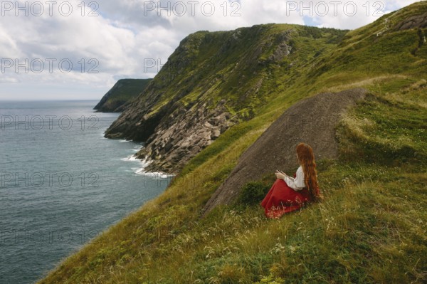Side view of an unrecognizable redhead woman in a red dress, seated on a grassy slope reading a book, overlooking the rugged coastline of Middle Cove, Newfoundland & Labrador, Canada