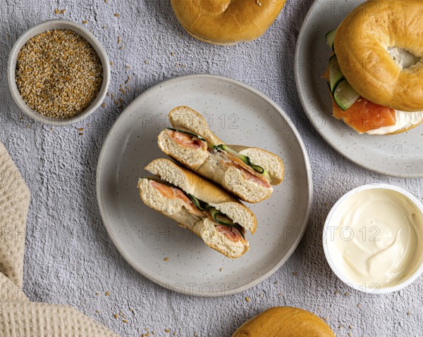 A top view of bagels with salmon and cream cheese on a speckled plate. A bowl of cream cheese and a bowl of seeds are nearby on a textured gray surface