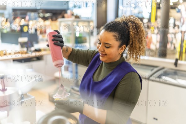 Woman employee in uniform smiling as she pours pink syrup over frozen yogurt in a cup at a shop counter, serving a fresh sweet treat to a customer in a casual small business setting