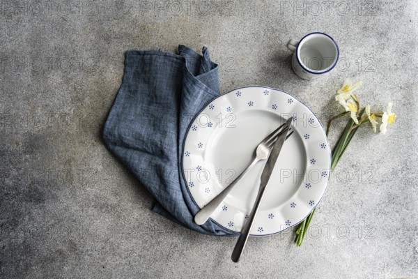 From above view of a stylish table setting featuring a white ceramic plate with blue dots, silver cutlery, a blue napkin, a small white cup, and white daffodils, all set on a textured grey surface