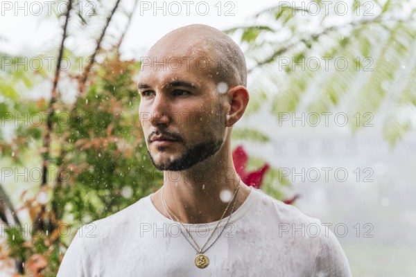 A man stands in the rain, deep in thought, surrounded by lush foliage in a forest. Drops fall around him, creating a contemplative and serene atmosphere in a natural setting