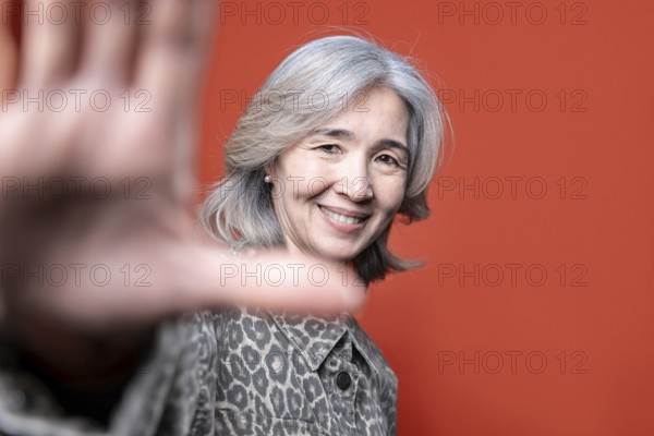 Asian senior woman smiles warmly while posing in front of a vibrant orange background. Her gray hair and patterned jacket add character, conveying confidence and positivity