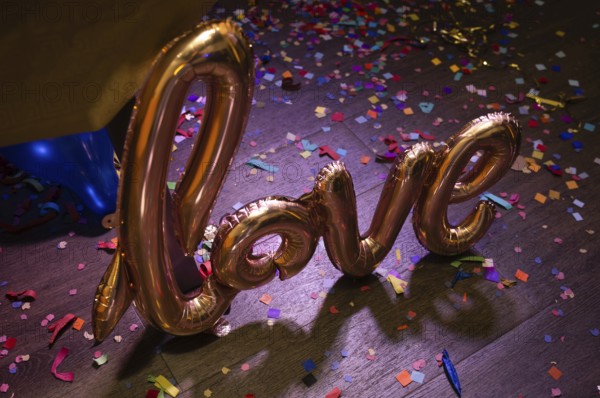 A golden foil balloon spelling love on a wooden floor covered in colorful confetti, creating a festive and romantic atmosphere perfect for celebrations
