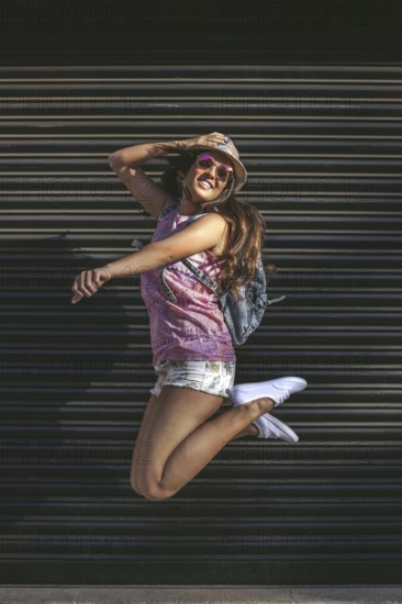 A young woman in sunglasses and a hat jumps joyfully against a dark urban background, embodying freedom and happiness. Perfect for lifestyle and youth-related projects