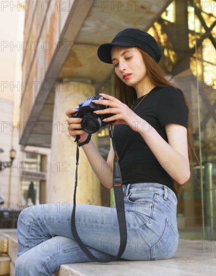 A woman photographer sits on a city bench, focused on her camera. Dressed in casual attire with a black cap, her thoughtful expression highlights her passion for photography