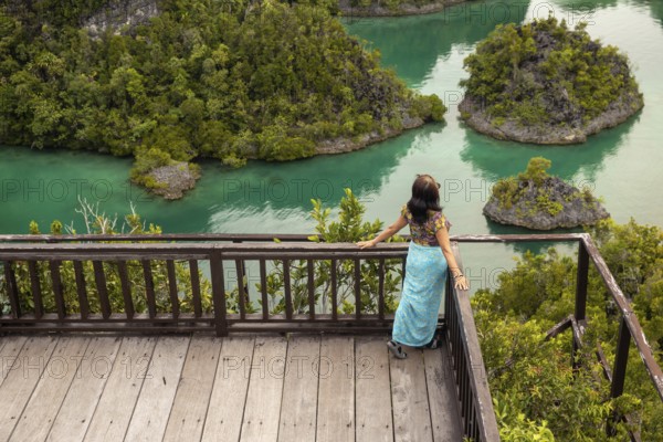 A woman stands on a wooden deck, admiring a lush tropical landscape with vibrant green water and islands of Indonesia. The peaceful scenery provides a sense of tranquility and escape