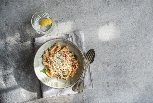 A delectable plate of cavatappi pasta topped with fresh basil, cheese and spicy tomato sauce accompanied by a glass of water with lemon, resting on a folded napkin with utensils