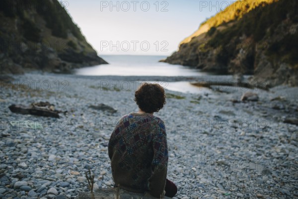 Back view of a man sitting contemplatively on a pebble beach, facing the tranquil sea as the sun sets behind the cliffs of Shoe Cove Beach