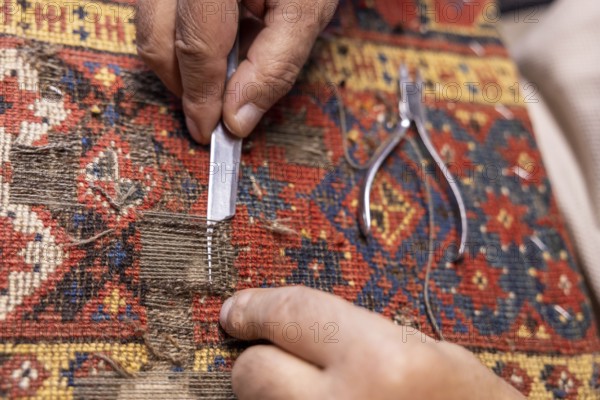 Close-up of a cropped unrecognizable senior male artisan's hands precisely restoring a colorful, traditional carpet using tools in his workshop in Salzburg