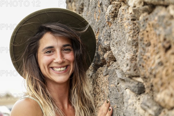 A woman wearing a hat smiles brightly while standing against a textured stone wall Her natural, carefree expression captures the essence of joyful outdoor adventures