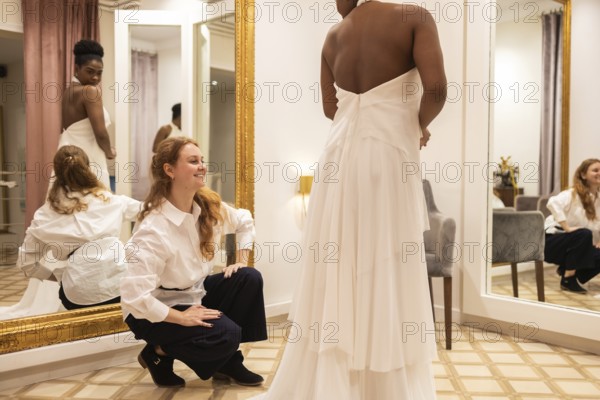 A bride to be wears a layered white gown, standing in a fitting room. Her designer, in a white shirt, assists her, ensuring a perfect fit. The room has elegant decor