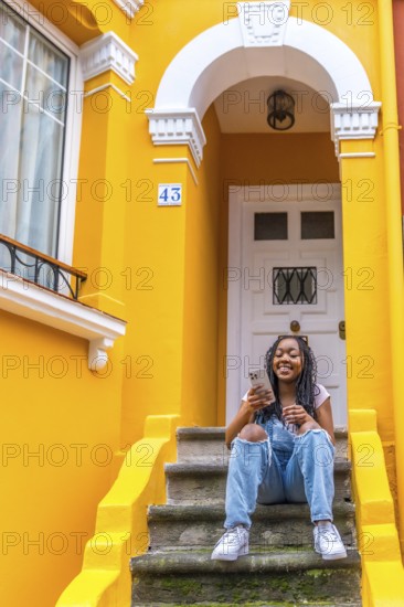 Vertical portrait of a smiling african woman using phone sitting in outside stairs of a yellow colored house