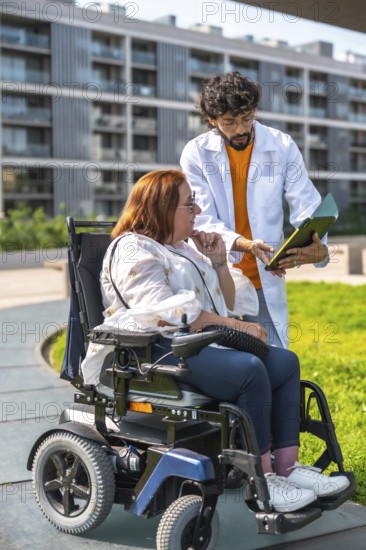 Doctor showing medical information on clipboard to woman using electric wheelchair outdoors in a hospital park