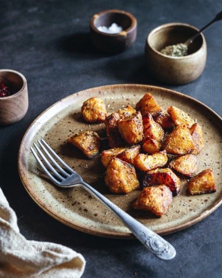 A rustic ceramic plate filled with crispy, herb roasted potatoes sits on a dark table. A fork and small wooden spice bowls accompany the dish, creating a cozy setting