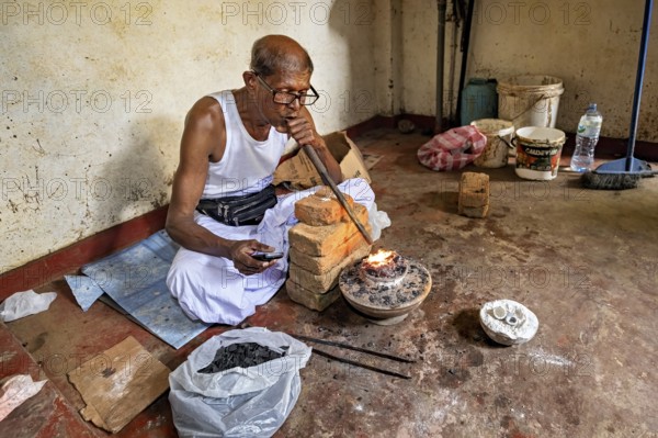 A man sits in a workshop, uses a bellows to start a fire, a man burns sapphires in a small oven in the town of Ratnapura in Sri Lanka