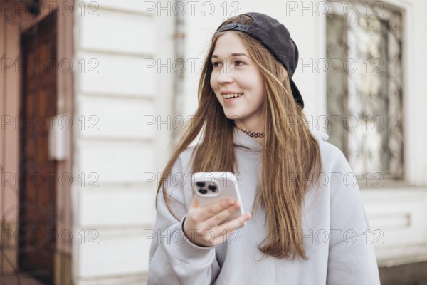 Teenage girl in casual attire with a hoodie and cap, smiling while holding a smartphone outdoors. The background shows an urban setting with a blurred building