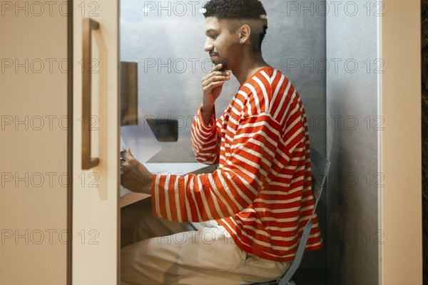 A man wearing a striped sweater works intently in a compact coworking phone booth, representing modern, flexible office solutions for productivity and concentration