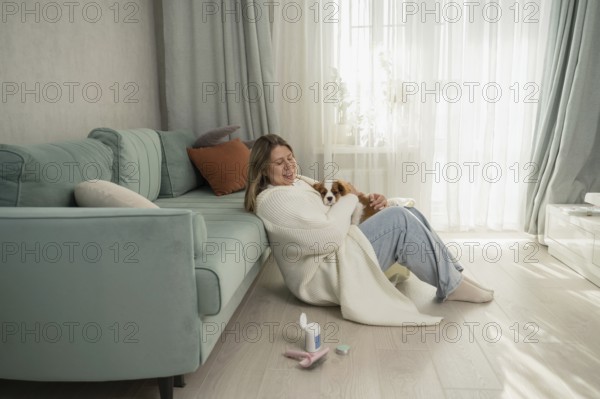 A woman cuddles her dog in a cozy home setting, sitting next to a teal sofa. Sunlight filters through sheer curtains, creating a peaceful and warm atmosphere