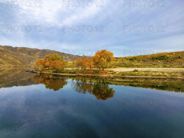 Aerial view capturing the serene beauty of Queenstown in autumn. The lake reflects the vibrant golden foliage and rugged hills, creating a breathtaking New Zealand landscape