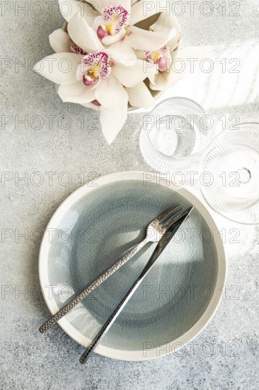 Stylish table setting features a textured ceramic plate, shiny cutlery, and delicate orchid flowers on a soft gray surface, creating a serene and elegant atmosphere