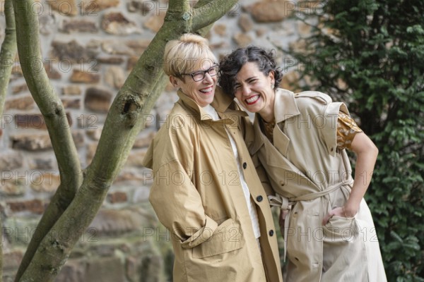 Two fashion women share a joyful moment outdoors, wearing casual trench coats. They laugh and lean on each other near a tree and a stone wall, embodying friendship and happiness
