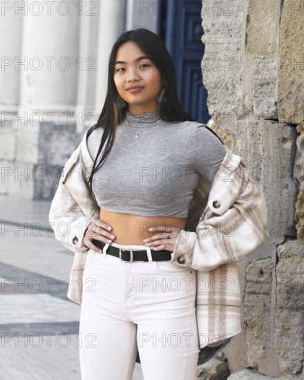 A young woman poses confidently in a chic urban setting, wearing a trendy plaid jacket over a gray crop top and high-waisted jeans, showcasing modern fashion and style