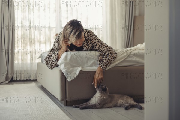 A woman in a stylish leopard-patterned robe leans over her bed to lovingly pet her cat in a cozy and well-lit bedroom, creating a serene moment of companionship