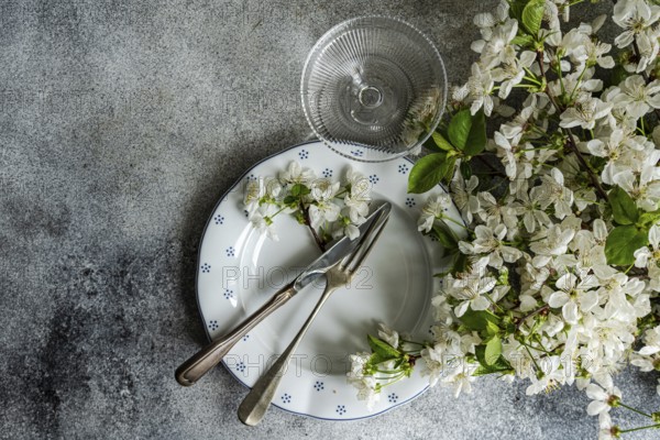 A table setting features vintage cutlery on a patterned plate, adorned with delicate white blossoms. A nearby crystal glass adds a touch of elegance to the rustic scene