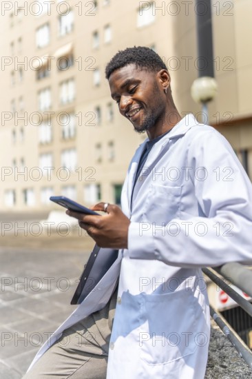 Vertical photo of a cheerful young african doctor using phone during work break outside the hospital