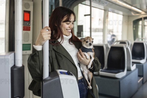 Smiling woman holding her chihuahua while riding on a tram. She wears a green coat and enjoys the journey. The tram interior is bright and modern, providing a comfortable commute