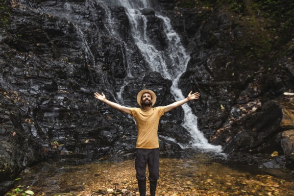 A young man stands with outstretched arms in front of a waterfall in the Choco Andino rainforest in Ecuador, embodying themes of adventure, ecotourism, and the profound connection with lush tropical landscapes