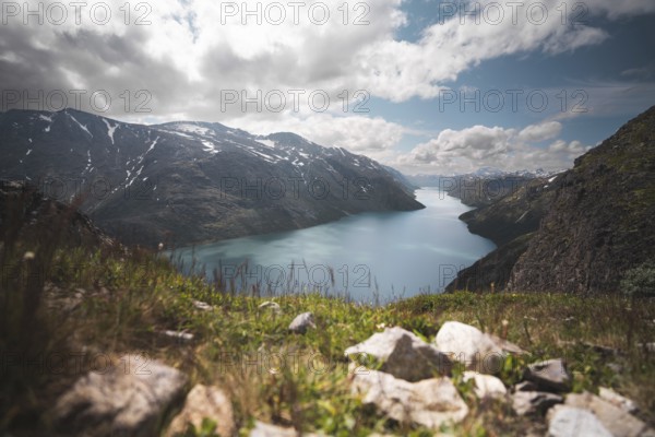 A breathtaking view of a serene turquoise lake nestled between rugged mountains in Norway The foreground features wild grass and rocks, adding depth to the dramatic landscape