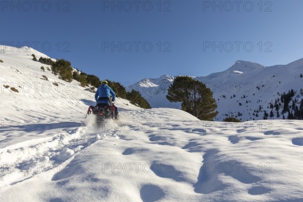 A man rides a snowmobile through a stunning winter mountain landscape, leaving a trail in the fresh snow under a clear blue sky. Evergreen trees and snow-capped peaks frame the scene