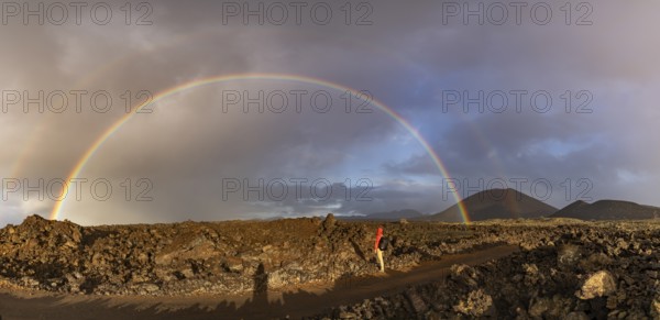 A vibrant rainbow arches over the rugged volcanic terrain of Timanfaya National Park in Lanzarote, Canary Islands, with a solitary figure standing in awe of the natural beauty