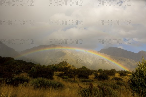 A breathtaking autumn scene at Mount Cook, New Zealand, featuring a vibrant rainbow arching over rugged mountains and lush greenery, under a dramatic cloudy sky