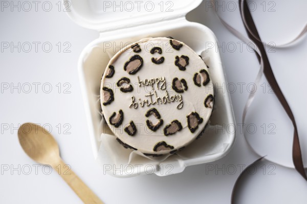 Top view of a mini bento birthday cake styled with a leopard print design and a Happy Birthday message, in a white takeout box, accompanied by a wooden spoon and satin ribbon