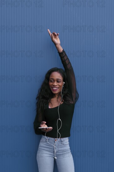 A young woman in casual attire, listening to music with earphones, raises her hand in excitement. She stands against a vibrant blue background, carrying a smartphone