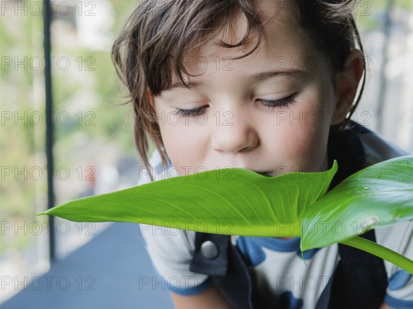 A young girl with tousled hair gently smells a large, shiny green leaf, capturing a moment of curiosity and interaction with nature