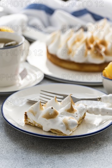 A slice of homemade lemon cake with toasted peaks on a plate, paired with a cup of tea. The scene captures a cozy dessert moment perfect for leisure or gathering